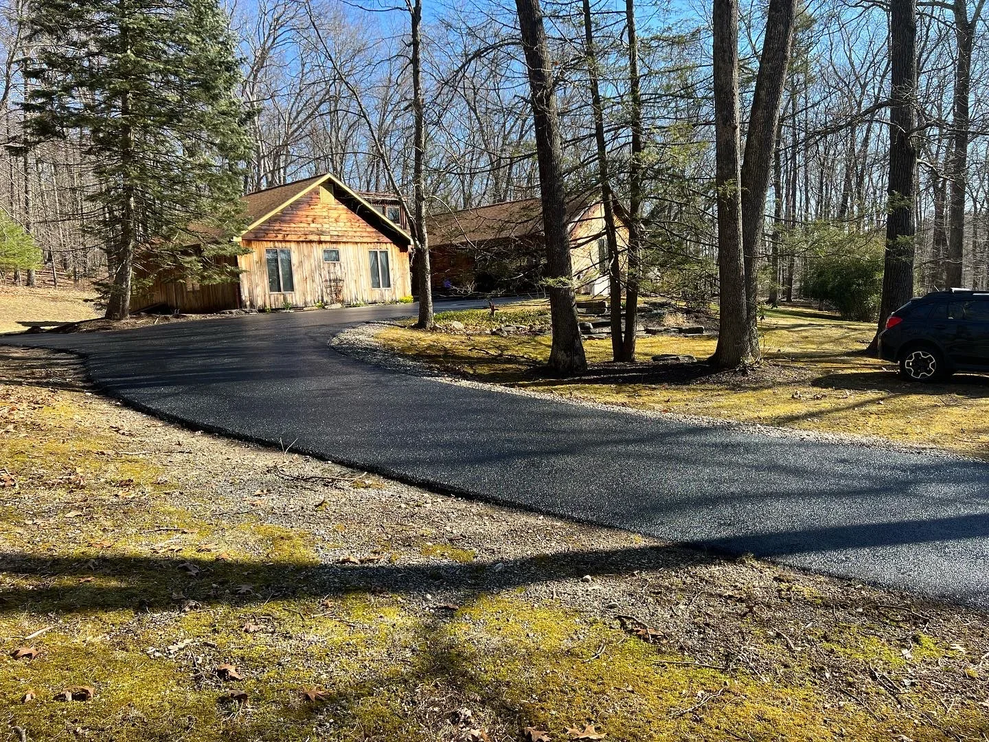 Newly paved asphalt driveway with curved design, gravel edges, residential home and tall trees