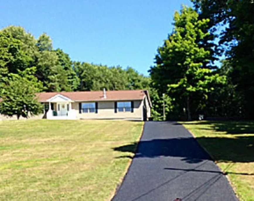 New black asphalt driveway leading to single-story home with white trim and brown roof