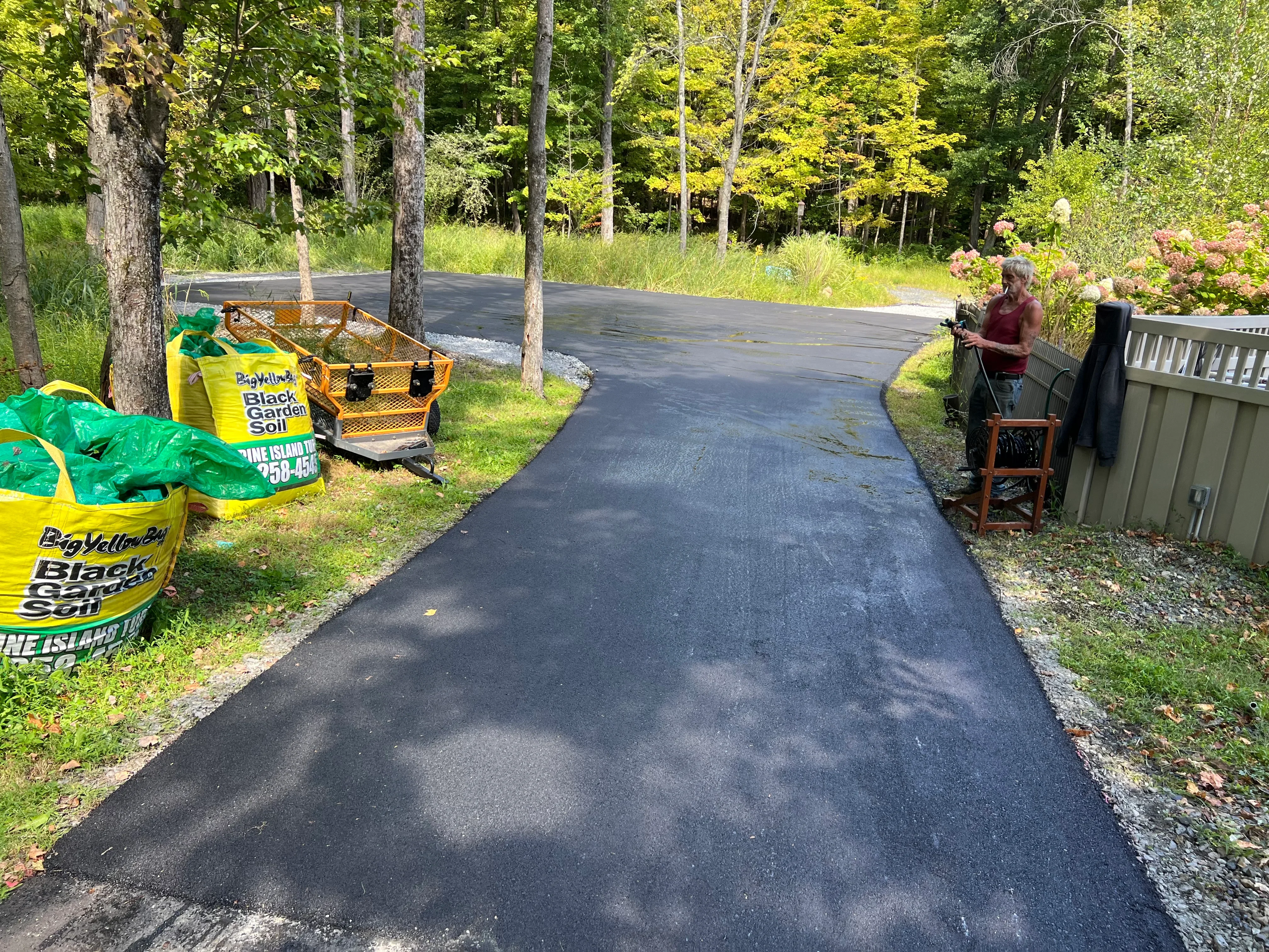 New asphalt driveway with dark surface, gravel edges, wooded setting with mature trees