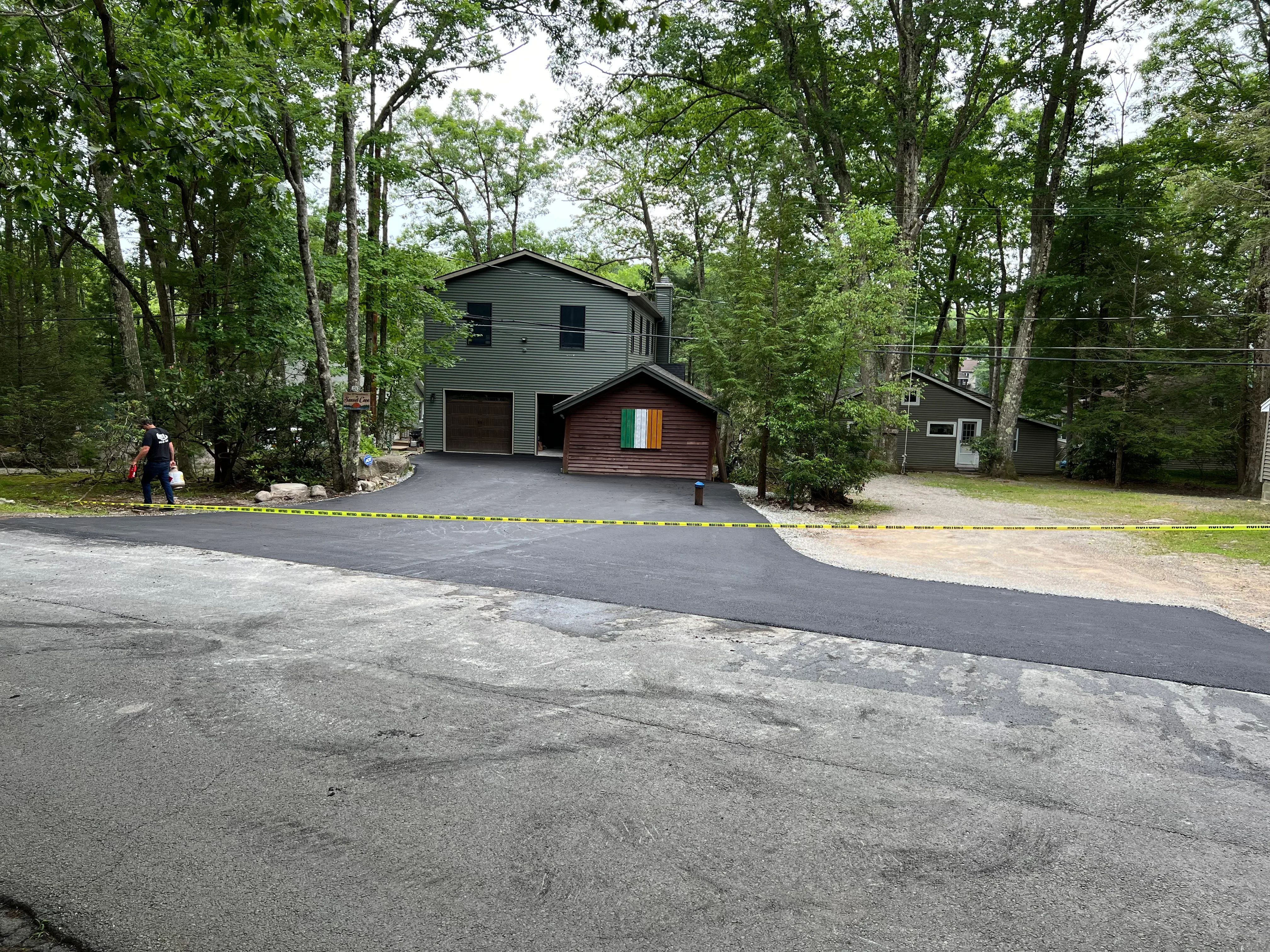 New black asphalt driveway with smooth finish leading to two-car garage on wooded residential lot