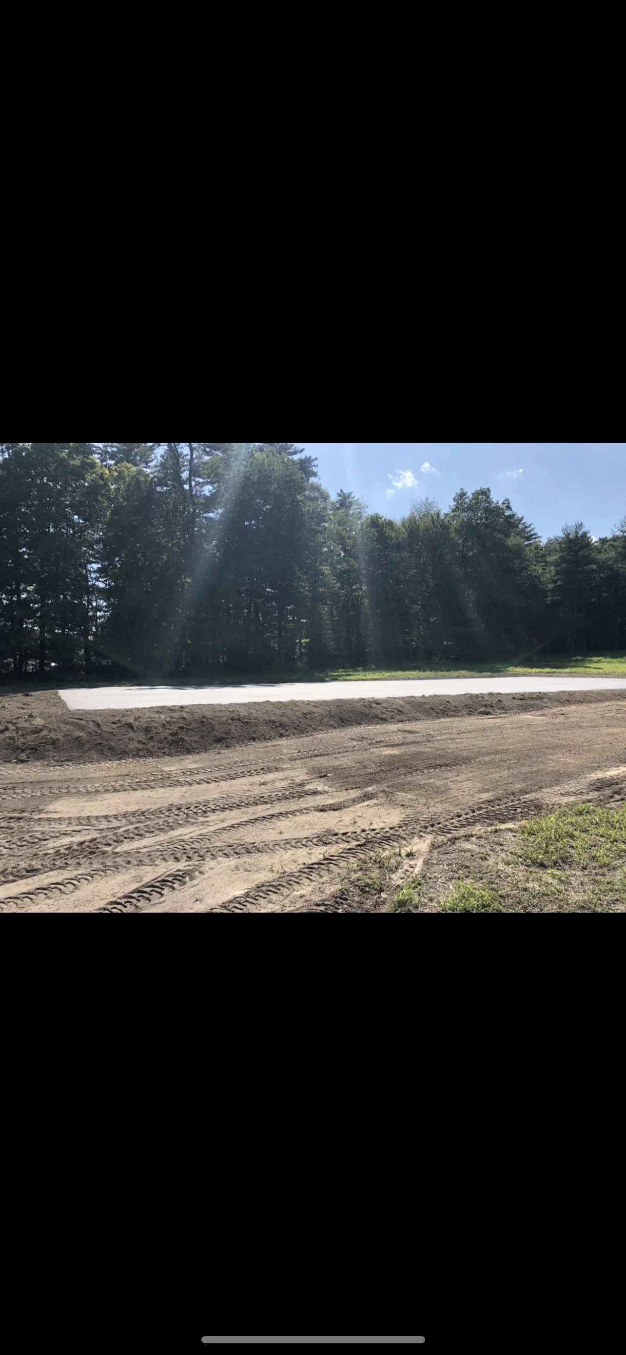 Excavated parking lot with compacted base, graded soil, and tree line backdrop
