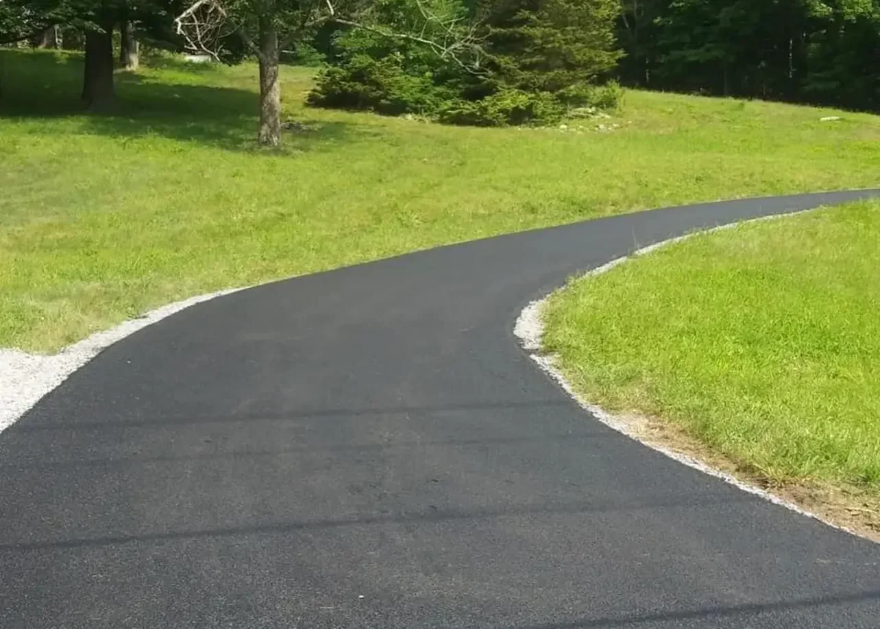 Curved asphalt driveway with white gravel border on residential property with trees and lawn