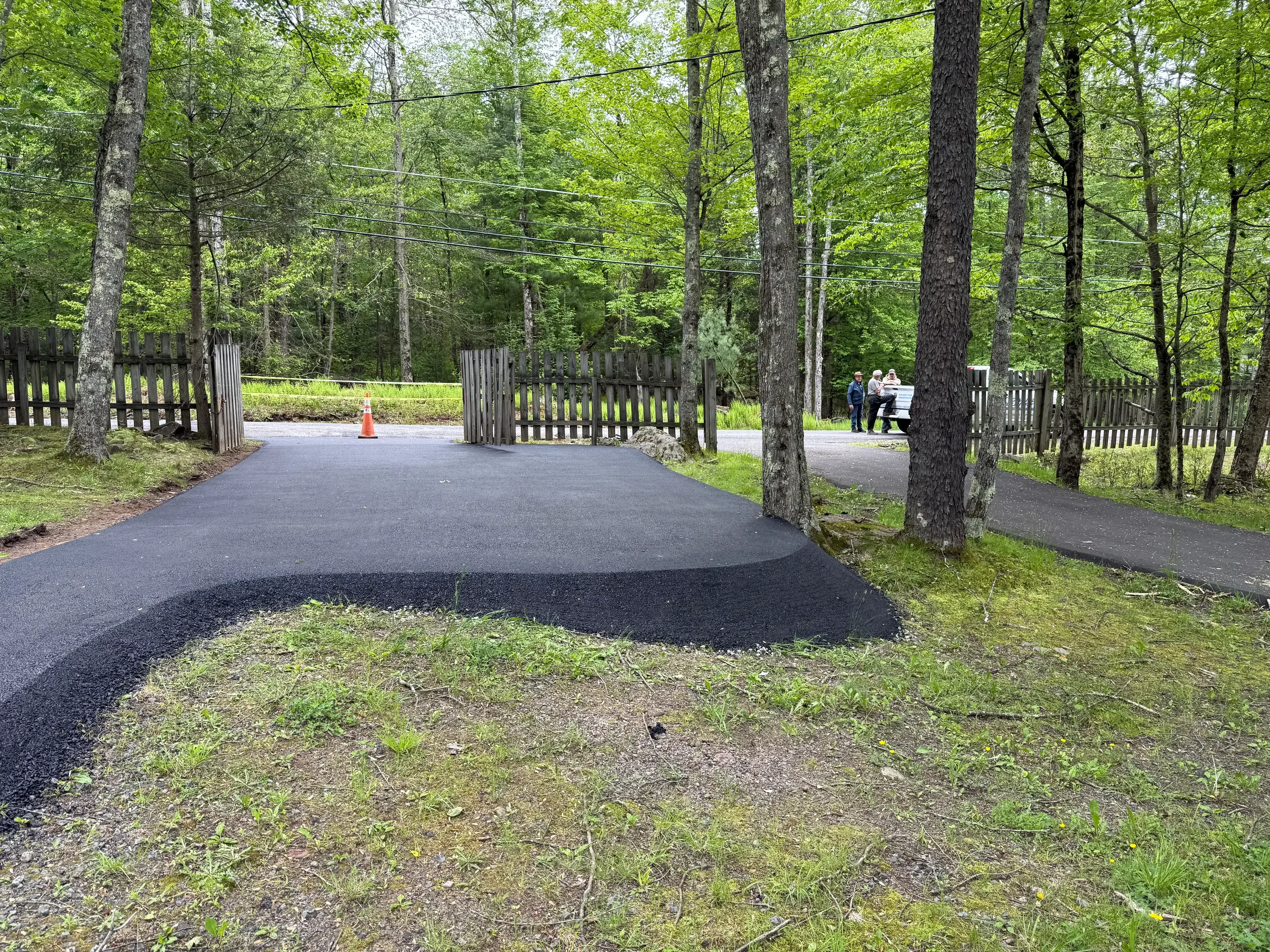 Curved asphalt driveway with white wooden gate and fence, surrounded by mature trees